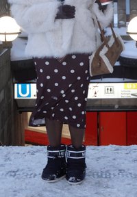 Personne portant un manteau en fourrure blanche, une jupe marron à pois, des collants noirs, des bottes d'hiver noires, tenant un sac à main marron doublé de fourrure et une tasse de café.