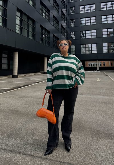Persona con suéter de rayas verdes y blancas, pantalones negros, gafas de sol blancas, sosteniendo un bolso naranja, parada en un área de estacionamiento urbana vacía.