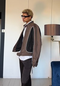 Νέος με ξανθά σγουρά μαλλιά, που φοράει μαύρα γυαλιά με πλαίσιο και ένα καφέ και μαύρο μπουφάν πάνω από λευκό πουκάμισο, στέκεται δίπλα σε έναν φωτιστικό δαπέδου.