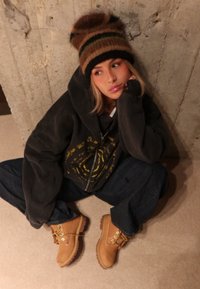 Stivali in pelle marrone con lacci gialli, felpa nera larga con disegno grafico, jeans blu scuro e berretto lavorato a maglia multicolore.