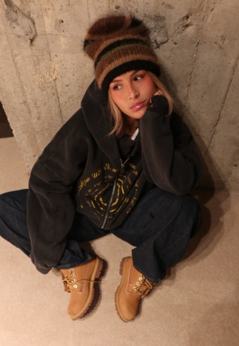 Bottes en cuir marron avec lacets jaunes, sweat à capuche noir ample avec motif graphique, jean bleu foncé et bonnet tricoté multicolore.