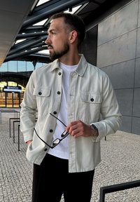 Gaisni pelēks, ar pogām aizdarāms žakets ar divām krūšpockets, uzvilkts virs baltas T-krekla. Persona tur brillēs un uz rokas ir pulkstenis.