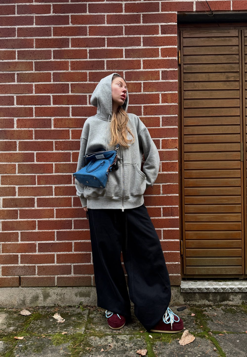 Sweat à capuche gris zippé avec une poche avant, associé à un pantalon noir ample. Sac à main bleu de forme distinctive, chaussures bordeaux avec des lacets bleu clair.
