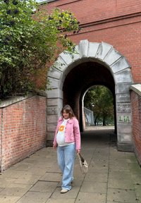 Zwangere vrouw in een roze jas en blauwe jeans die met een beige handtas wandelt naast een bakstenen boog en groenvoorzieningen.