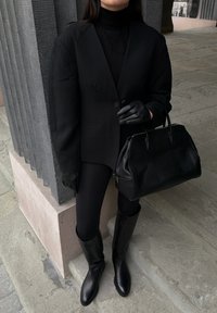 Sort oversize blazer, rullekrave og leggings kombineret med knælange sorte læderstøvler. Sort håndtaske med to håndtag og tekstureret overflade.