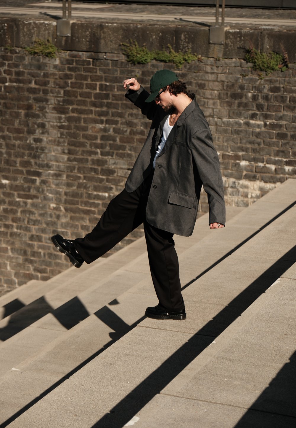 Persona, kas valkā pelēku pārāk lielu žaketu, baltu topiņu un melnus bikses, dodas lejā pa betona kāpnēm, ar zaļu cepuri un melnām kurpēm.