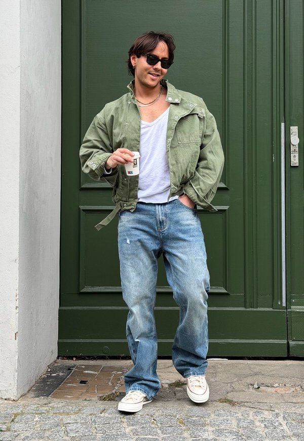 Hombre con gafas de sol, chaqueta verde, camisa blanca, vaqueros azules y zapatillas blancas sosteniendo una pequeña taza de café, de pie frente a una puerta verde.