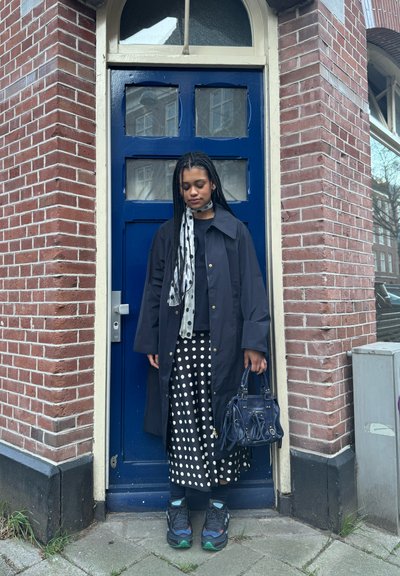 Abrigo impermeable negro sobre una parte superior negra, falda con lunares blancos, accessorios con una bufanda estampada y un bolso azul marino. Zapatillas negras.