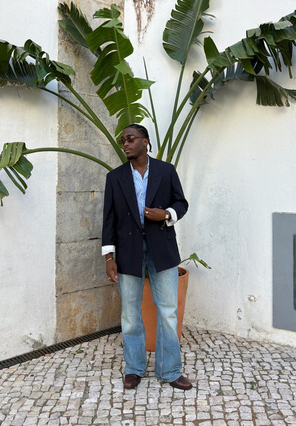 Hombre con gafas de sol que lleva una chaqueta oscura, camisa azul a rayas, vaqueros y zapatos marrones, de pie sobre adoquines junto a una gran planta verde en maceta.