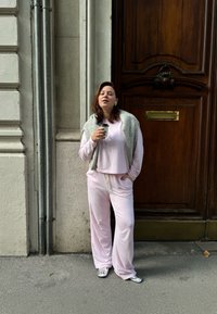 Vaaleanpunainen vapaa-ajan asu, jossa on väljä malli yläosassa ja housuissa, yhdistettynä harmaaseen neuleeseen, joka on heitetty olkapäiden yli, sekä metallinvärisiin kenkiin. Käsissä kahvimuki.