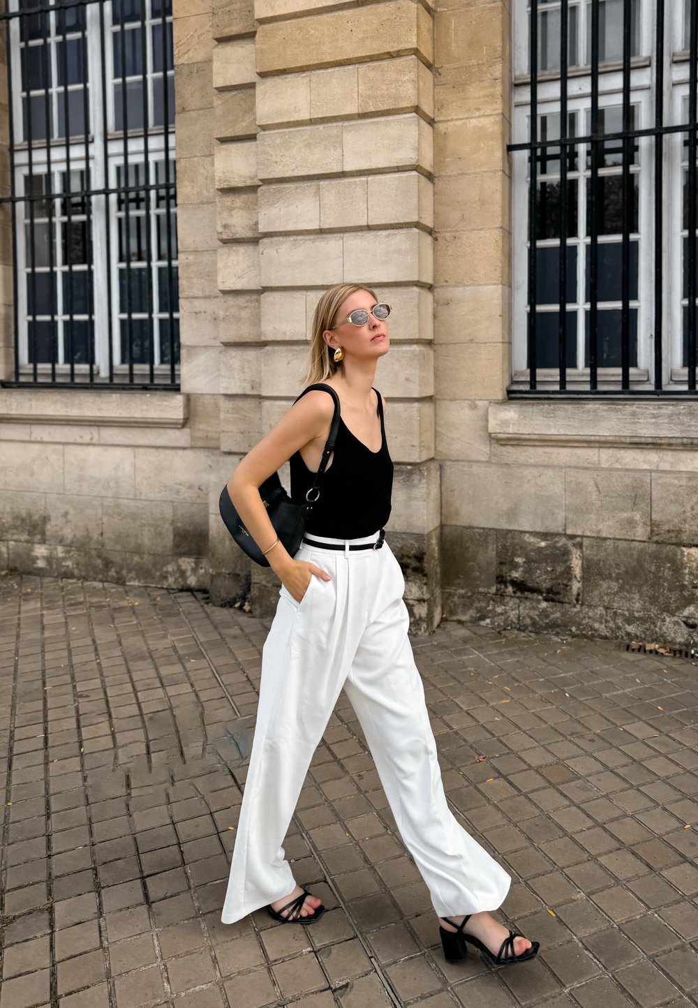 Schwarzes ärmelloses Top, weiße weit geschnittene Hose und schwarze Sandalen. Die Accessoires umfassen große kreisförmige Ohrringe und eine Sonnenbrille. Trägt eine schwarze Tasche.