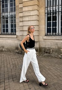 Schwarzes ärmelloses Top, weiße weit geschnittene Hose und schwarze Sandalen. Die Accessoires umfassen große kreisförmige Ohrringe und eine Sonnenbrille. Trägt eine schwarze Tasche.