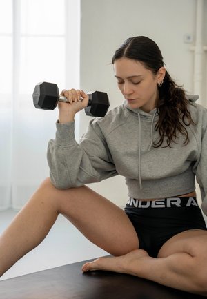 Mujer con sudadera con capucha gris y pantalones cortos negros sentada con las piernas cruzadas, levantando una mancuerna hexagonal negra durante un entrenamiento en interiores.