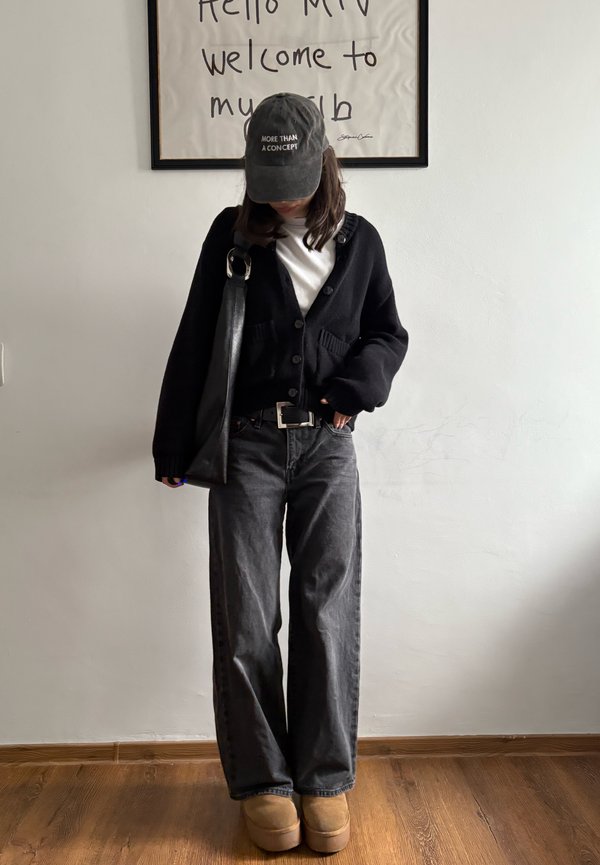 Personne portant un cardigan noir, un jean gris à jambes larges, des bottes marron clair et une casquette noire avec le texte « PLUS QU'UN CONCEPT », debout sous une œuvre encadrée accrochée au mur.