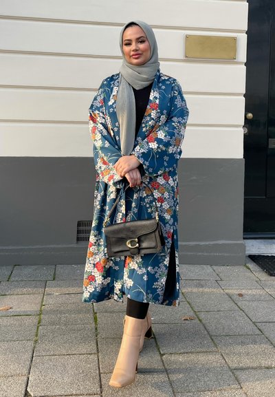 Kimono floral largo de tela azul con flores multicolores, combinado con leggings negros y botines de tacón bloque en nude; sostiene un bolso negro.