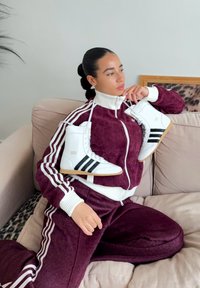 Femme en survêtement Adidas bordeaux assise sur un canapé beige, tenant une paire de baskets hautes blanches Adidas sur ses épaules.