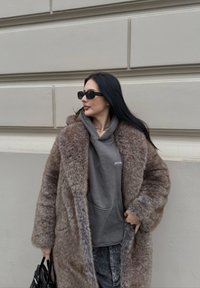 Καφέ ψεύτικο γούνα παλτό πάνω από γκρι φούτερ με κουκούλα, με πλούσια υφή, χαλαρή εφαρμογή και μινιμαλιστικές λεπτομέρειες; συνοδευόμενο από μαύρα γυαλιά ηλίου.