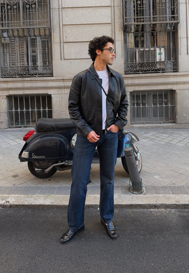 Homme portant une veste en cuir noire, une chemise blanche, un jean bleu et des lunettes de soleil, se tenant sur le trottoir près d'un scooter Vespa noir garé.