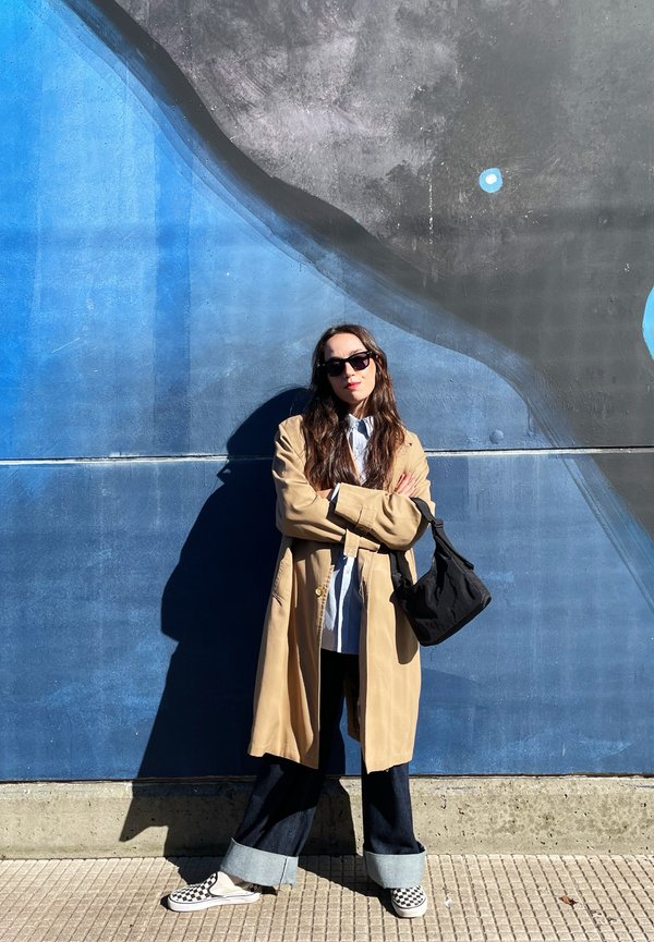 Cappotto trench beige sopra una camicia bianca, jeans larghi scuri con orli risvoltati, e scarpe slip-on a scacchi. Borsa a tracolla nera e occhiali da sole.