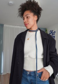 Lungo cappotto scuro sopra un maglione bianco strutturato e jeans di denim. Una collana choker di denim impreziosisce il look. Stanza con tende di colore chiaro sullo sfondo.