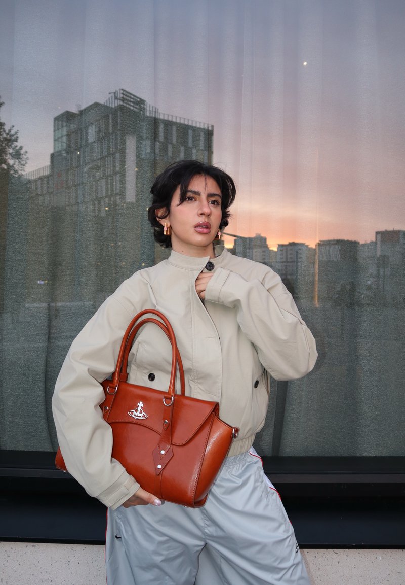 Jeune personne en veste beige et pantalon clair tient un sac à main en cuir marron, debout devant une fenêtre reflétant des bâtiments de la ville au coucher du soleil.