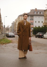 Manteau brun à double boutonnage, pantalon clair et cravate. Porte un sac brun. Scène urbaine avec voitures garées et bâtiments. Ciel couvert.