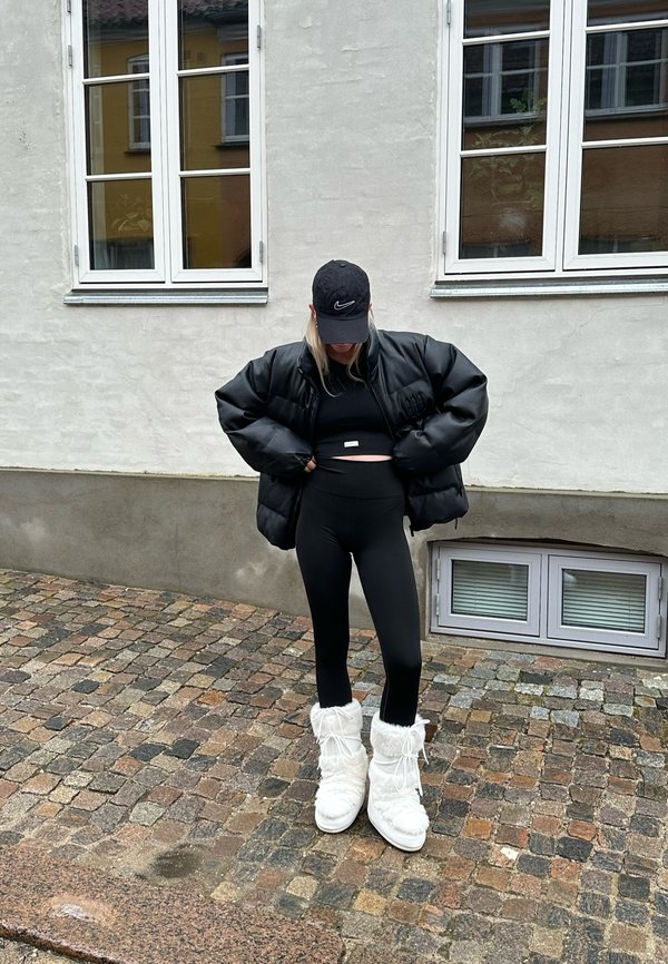 Czarna puchowa kurtka, czarny top crop, czarne legginsy z wysokim stanem i białe pluszowe botki z futrzanymi akcentami; na tle teksturowanej powierzchni brukowej.