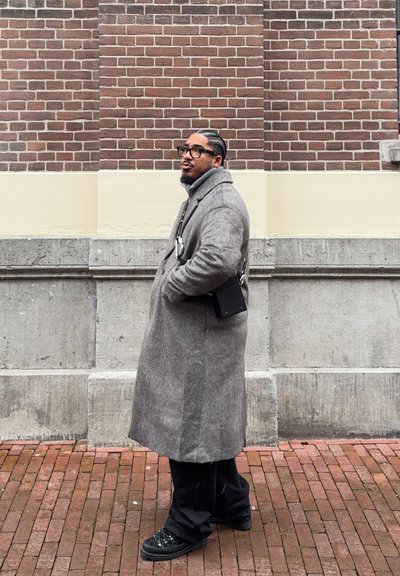 Hombre con cabello trenzado y gafas, vestido con un abrigo largo gris y zapatos negros, de pie sobre un pavimento de ladrillo frente a una pared de ladrillo y piedra.
