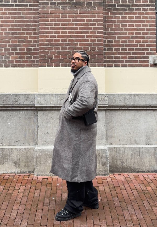 Hombre con cabello trenzado y gafas, vestido con un abrigo largo gris y zapatos negros, de pie sobre un pavimento de ladrillo frente a una pared de ladrillo y piedra.