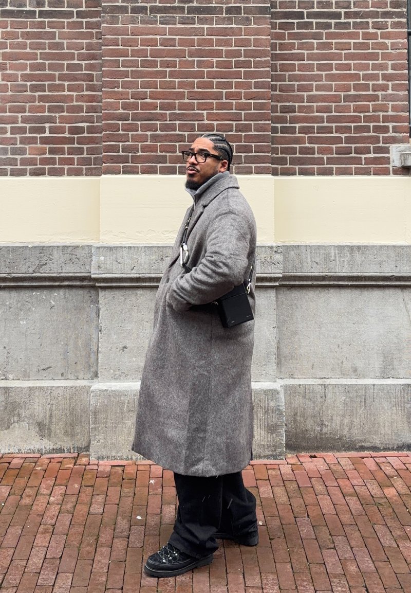 Homme aux cheveux tressés et portant des lunettes, vêtu d'un long manteau gris et de chaussures noires, debout sur un pavement en briques devant un mur en briques et en pierre.