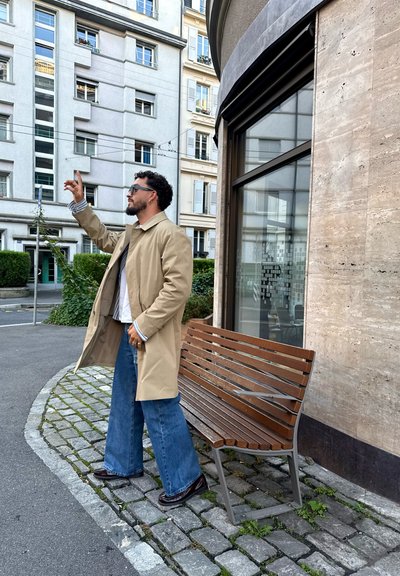 Hombre con abrigo beige y jeans azul de pie en una acera de adoquines cerca de un banco de madera, señalando hacia arriba con la mano levantada.