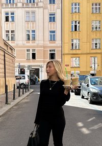 Čierna dlhá rukávová košeľa a nohavice, drží dva ľadové nápoje v priehľadných pohároch so zelenými slamkami, stojí na mestskom námestí s budovami za sebou.