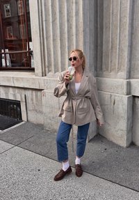 Beige oversized blazer met een gecinchde taille, gecombineerd met blauwe cropped jeans en bruine schoenen. Houdt een transparante drank vast en staat tegen een gestructureerde muur.