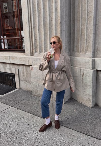 Blazer oversize beige con cintura ajustada, combinado con vaqueros cortos azules y zapatos marrones. Sosteniendo una bebida transparente, de pie contra una pared texturizada.