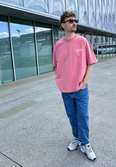 Camiseta de gran tamaño en rosa claro con el logo de "ALLSAINTS", combinada con jeans de mezclilla azul y zapatillas blancas. De fondo se ve un moderno edificio de cristal.