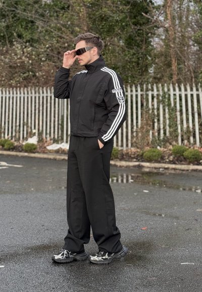Chaqueta deportiva negra con tres franjas blancas y logotipo, pantalones anchos negros y zapatillas metálicas. La persona ajusta unas gafas de sol elegantes.