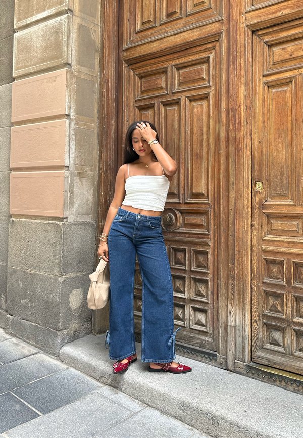 Valkoinen ribattua materiaalia oleva crop-top, siniset leveälahkeiset farkut solmuvuoroilla, tummanoranssia liukuvia tossuja, jotka on koristeltu, seisoo puisia ovia vasten.