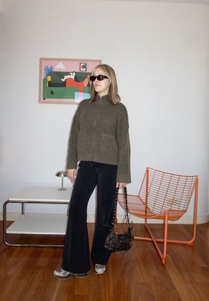 Mujer con gafas de sol, suéter verde oliva con cremallera, pantalones negros de pierna ancha, sosteniendo un bolso negro, de pie en el interior cerca de una silla de alambre naranja y una mesa auxiliar.