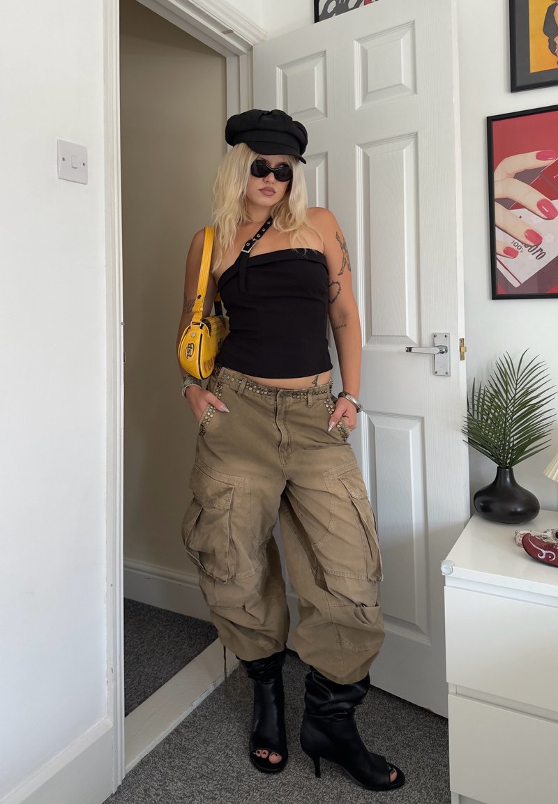 Femme portant un bustier noir, un chapeau noir, des lunettes de soleil, un pantalon cargo beige, des bottes noires à bout ouvert, et portant un sac à bandoulière jaune à l'intérieur.
