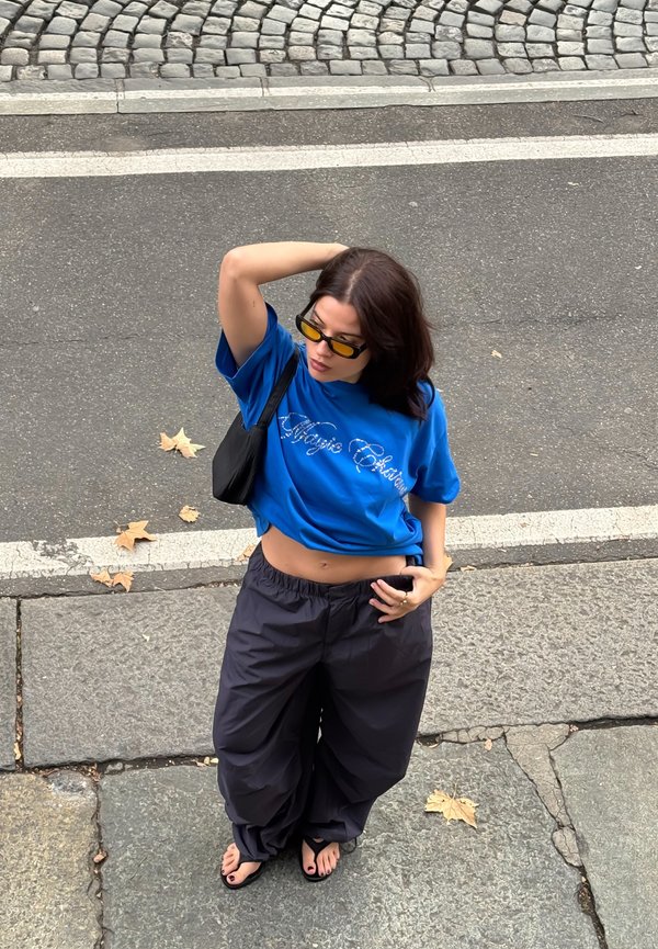 Une femme portant des lunettes de soleil aux verres teintés de jaune, un T-shirt bleu et un pantalon sombre ample pose dans une rue pavée, une main derrière la tête.