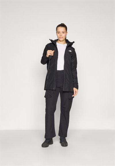 Chaqueta negra impermeable con cierre frontal, capucha ajustable y logotipo. Combinada con pantalones cargo negros y calzado negro. Textura suave.