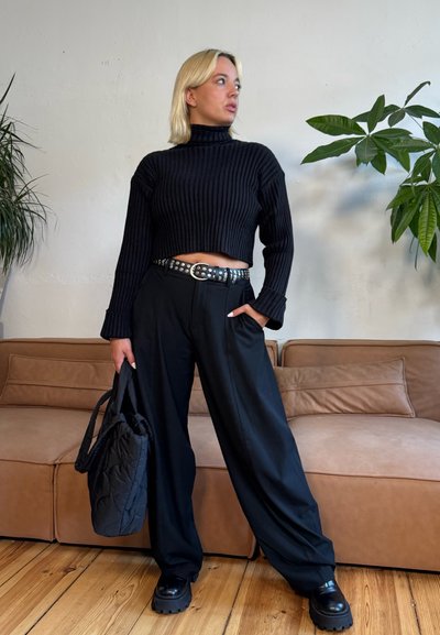 Suéter negro recortado de canalé con mangas largas, cuello alto, combinado con pantalones anchos negros, cinturón plateado con tachuelas y bolso negro acolchado.