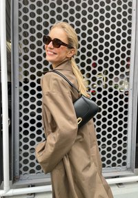 Hnedý prehnaný trenčkot, čierna kabelka so zlatým kovovým vybavením, slnečné okuliare s vzorom, textúrované pozadie s kruhovými perforáciami.