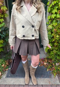 Light beige double-breasted jacket, brown pleated skirt, knee-high beige boots, and a brown woven handbag with textured accents.