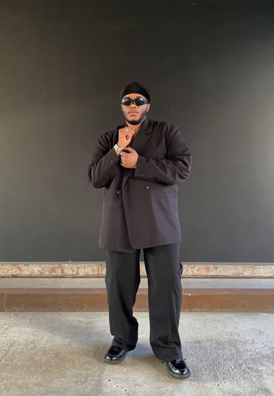 Blazer negro de doble botonadura sobre una camisa negra, combinado con pantalones negros. Gafas de sol negras y un reloj plateado completan el conjunto.