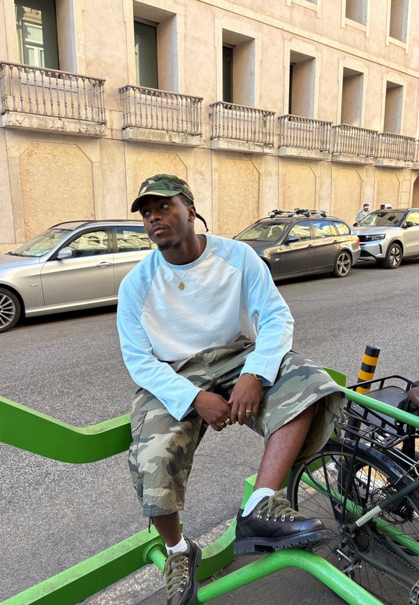 Hombre con gorra y pantalones de camuflaje sentado sobre un aparcabicicletas verde en una calle urbana con coches aparcados y un edificio beige al fondo.