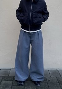 Veste aviateur marine avec col côtelé, ceinture rayée claire, associée à un pantalon gris large et ample. Chaussures noires à finition texturée.