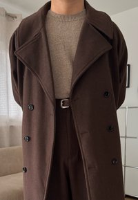 Osoba ubrana w brązowy dwurzędowy płaszcz na beżowym swetrze dzianym, ciemnobrązowe spodnie i brązowy skórzany pasek, wewnątrz pomieszczenia.