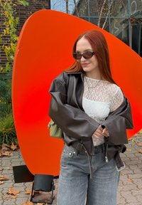 Rudos odos laisvo kirpimo striukė, balta nėriniuota palaidinė, pilki džinsai, žalia rankinė. Fone matyti didelė oranžinė abstrakti forma.