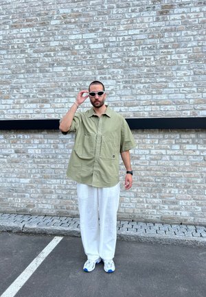 Camisa de botón extragrande color verde oliva con dos bolsillos en la parte frontal, combinada con pantalones de lino blancos y zapatillas blancas con acentos azules.